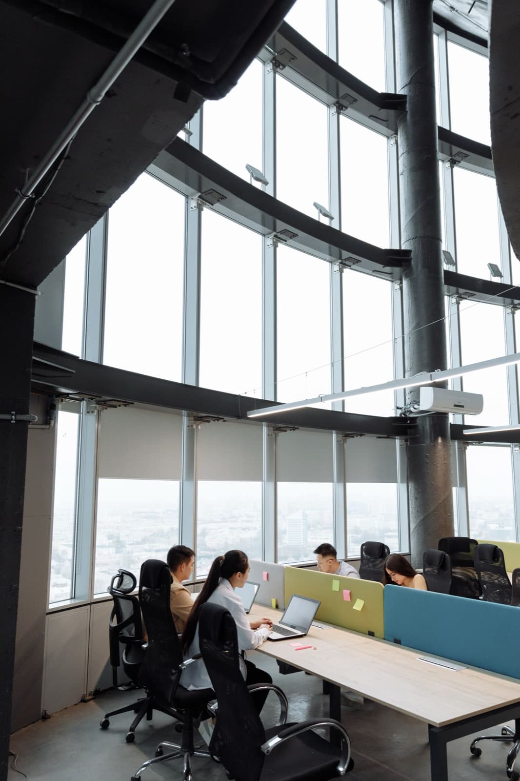 People working at long shared desks beside a wall of windows in a high-rise office.